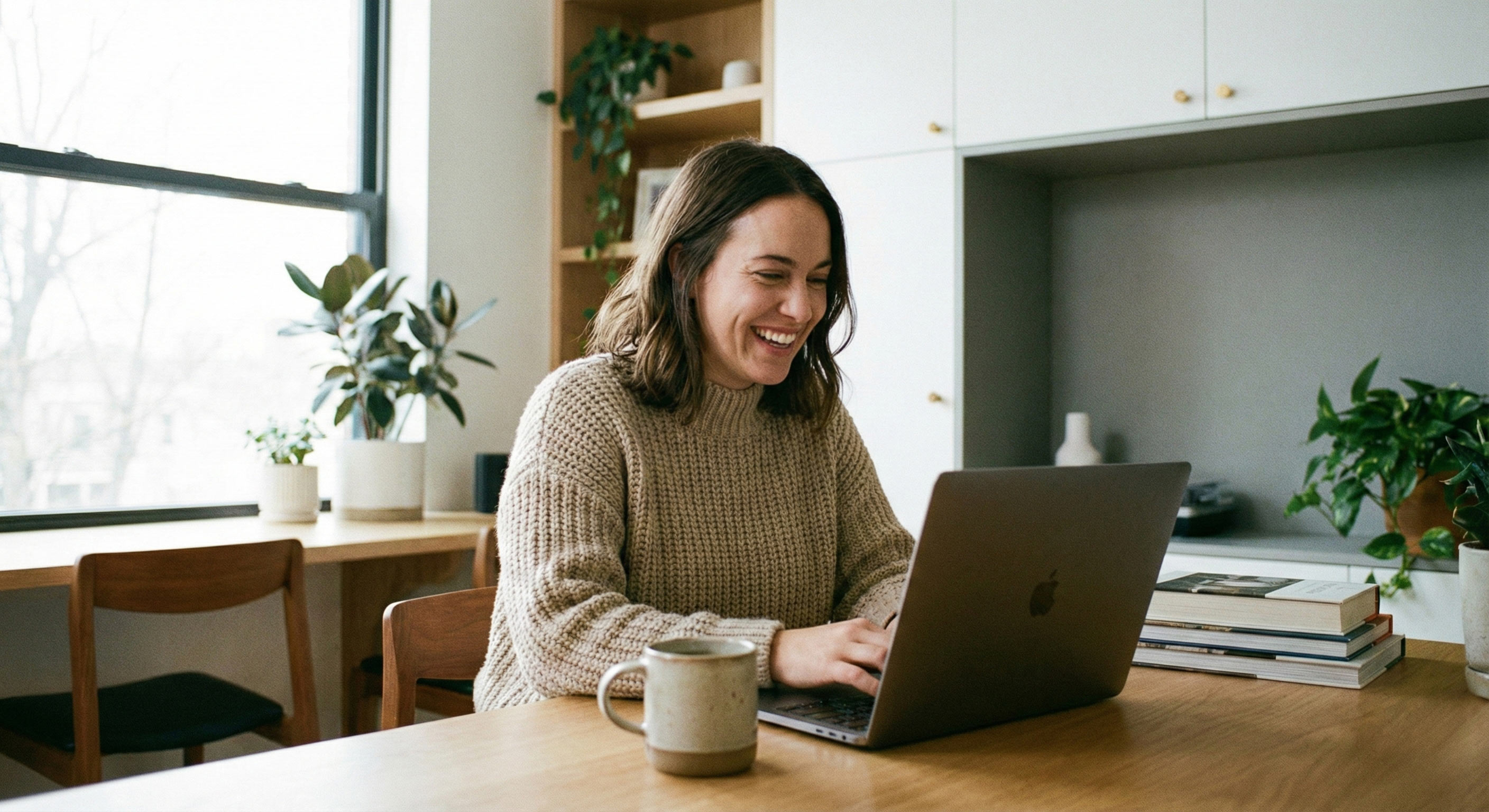 A medium shot of a woman in a modern home office, smiling while looking at a laptop screen, with a coffee mug on the desk.